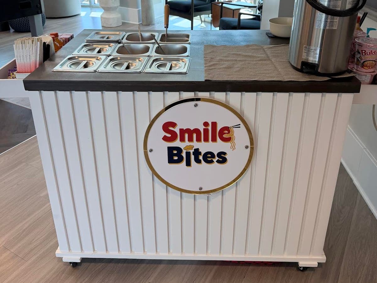 The SmileBites ramen cart, white panelled with a gold-rimmed logo medallion, set up indoors with nine stainless toppings wells along the counter