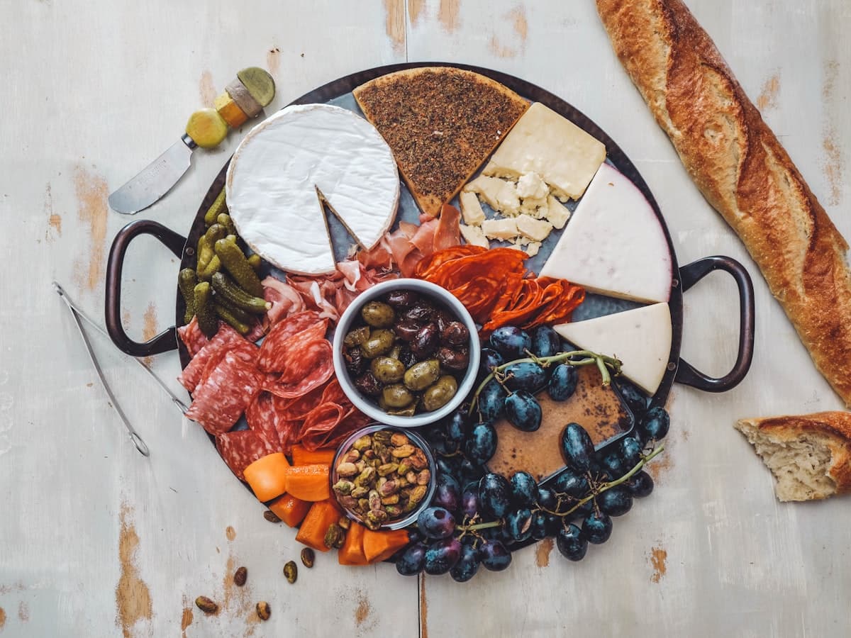 A round charcuterie board with cured meats, soft cheeses, olives, grapes and a sliced baguette