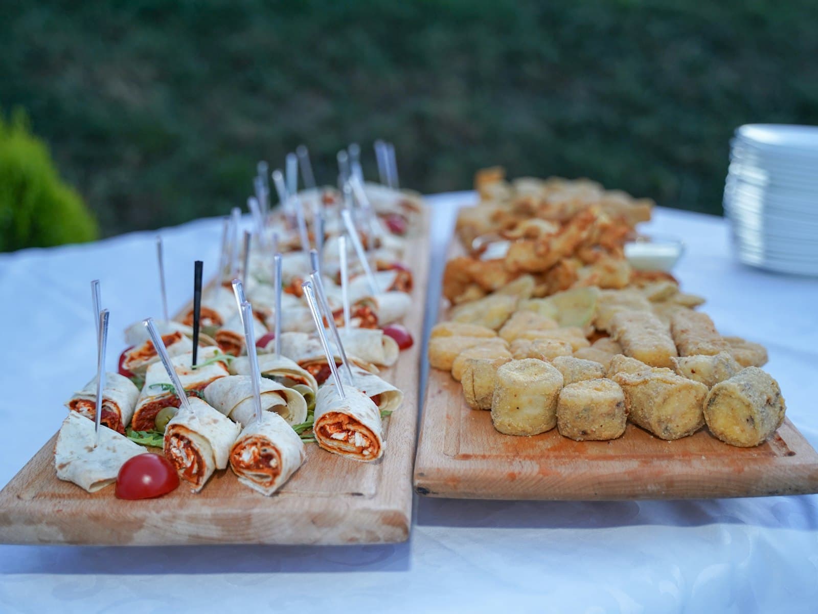 Wooden boards of rolled wraps on picks and breaded fried bites, set out on a white cloth outdoors