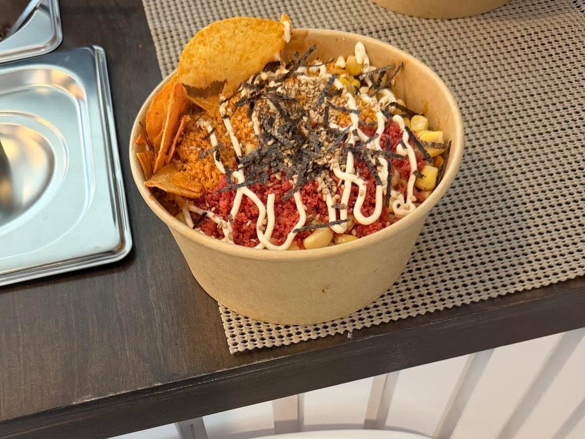 A kraft bowl of saucy ramen topped with Takis dust, furikake, nori, sweetcorn and a creamy mayo drizzle, with a bag of chips at the side